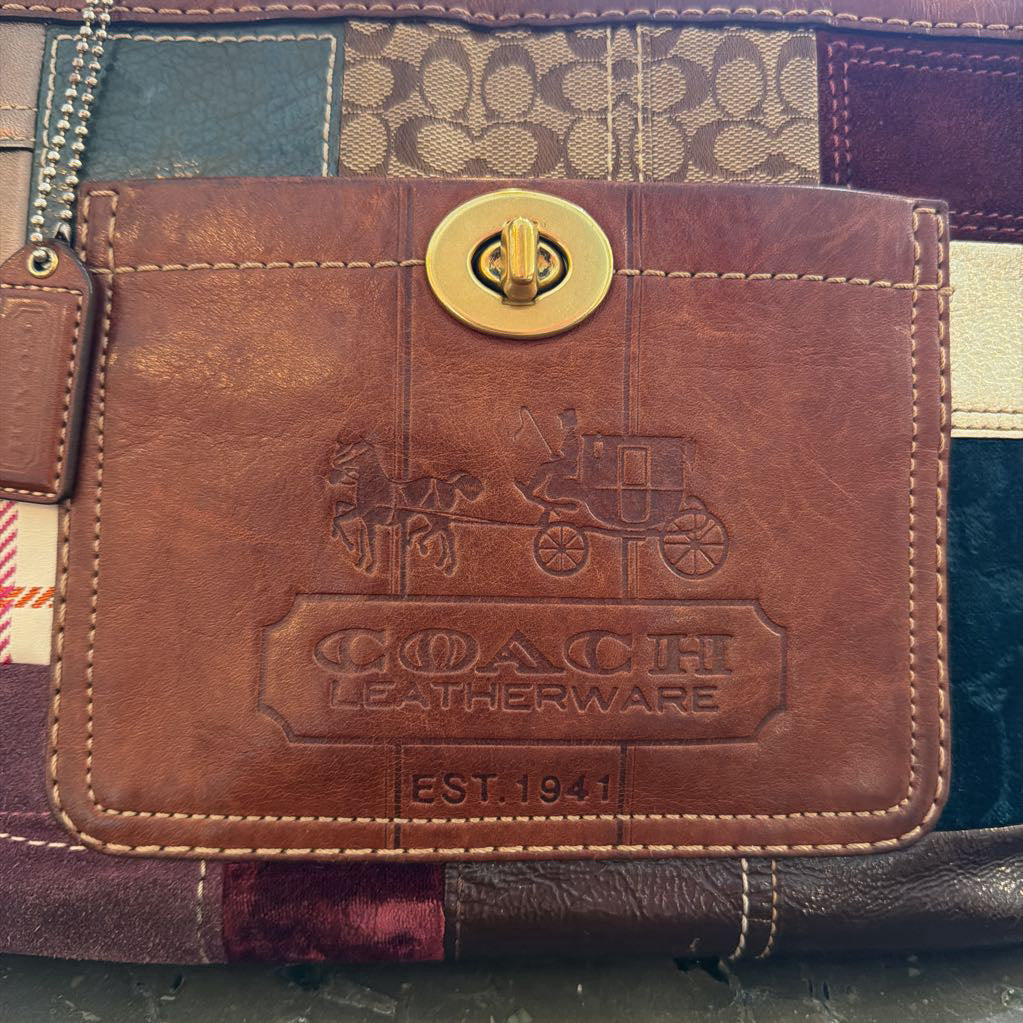 A close-up of a Coach leather wallet or card holder with a gold-tone turn-lock closure and embossed Coach Leatherware branding on the front, set against a patchwork patterned background.