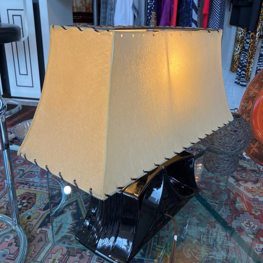 A vintage-style table lamp with a large beige fabric drum shade and a glossy black sculptural base, shown on a glass table in a cluttered antique store or home decor shop.