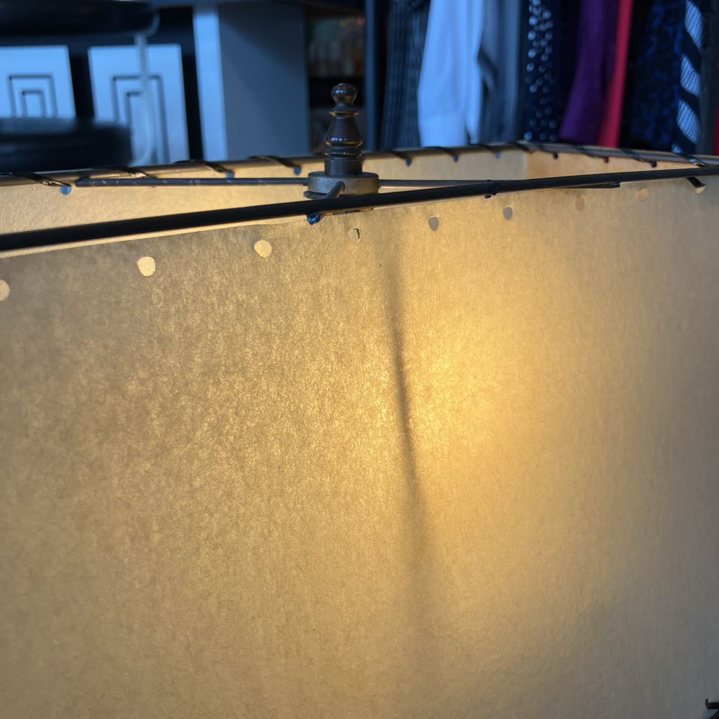A close-up of a lampshade with a warm, glowing yellow-gold light emanating from within. The top rim shows a black metal frame with brass-colored decorative finials and small white dots along the edge.