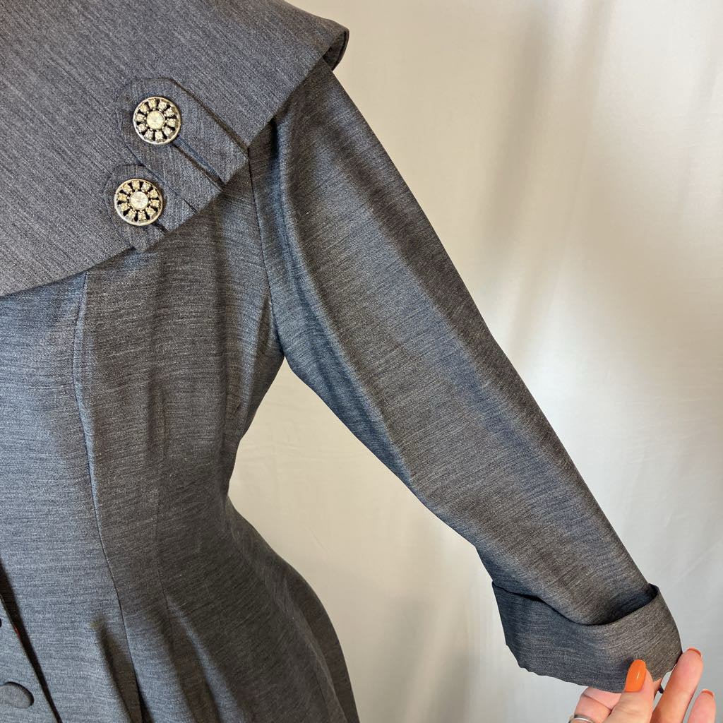 Close-up of a gray textured long-sleeve dress or coat with a wide collar and decorative round buttons, shown on a person against a plain light background.