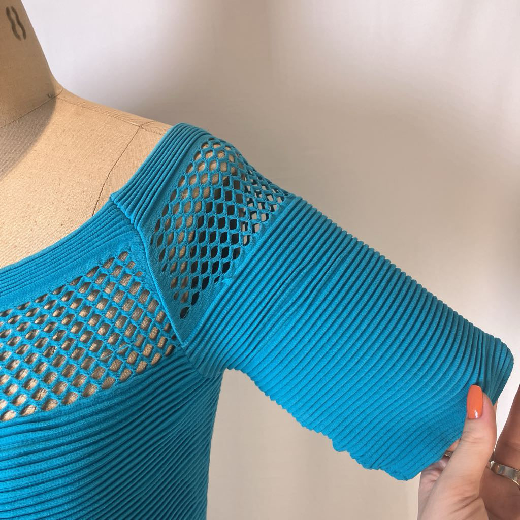 A bright turquoise blue ribbed knit top with a large fishnet cutout on the shoulder and sleeve, displayed on a beige dress form with a hand holding the sleeve.