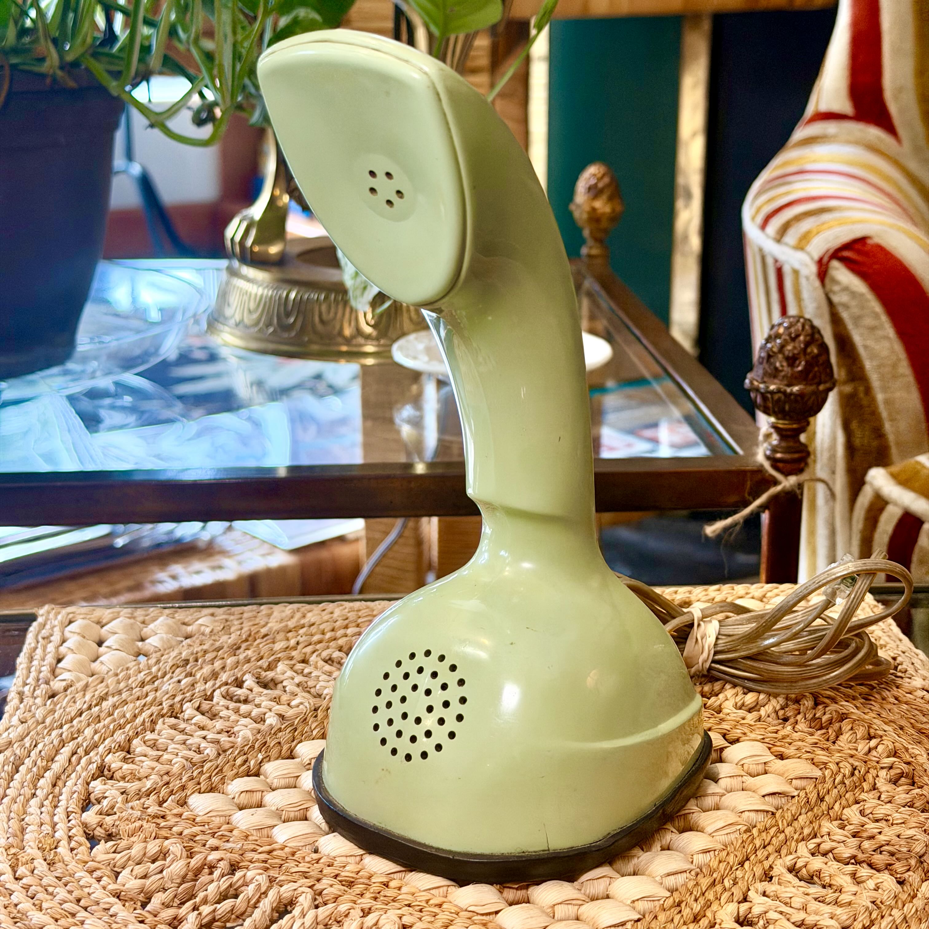 A vintage light green rotary telephone receiver handset resting on a woven placemat on a glass-top table, with a coiled cord nearby and an indoor room background.