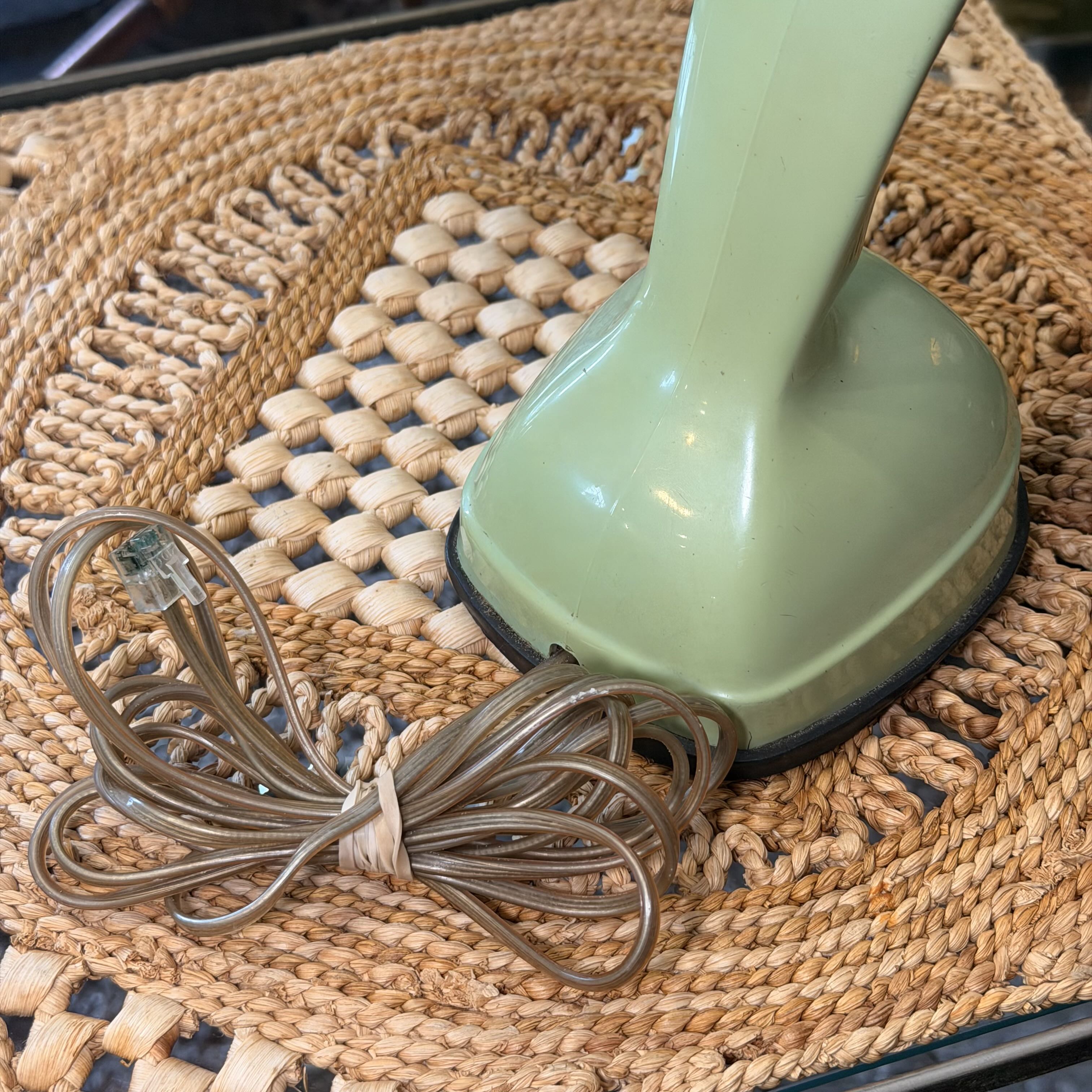 A vintage-style electric iron or heat styling tool with a pale sage green handle and a coiled brown cord, resting on a woven straw placemat.