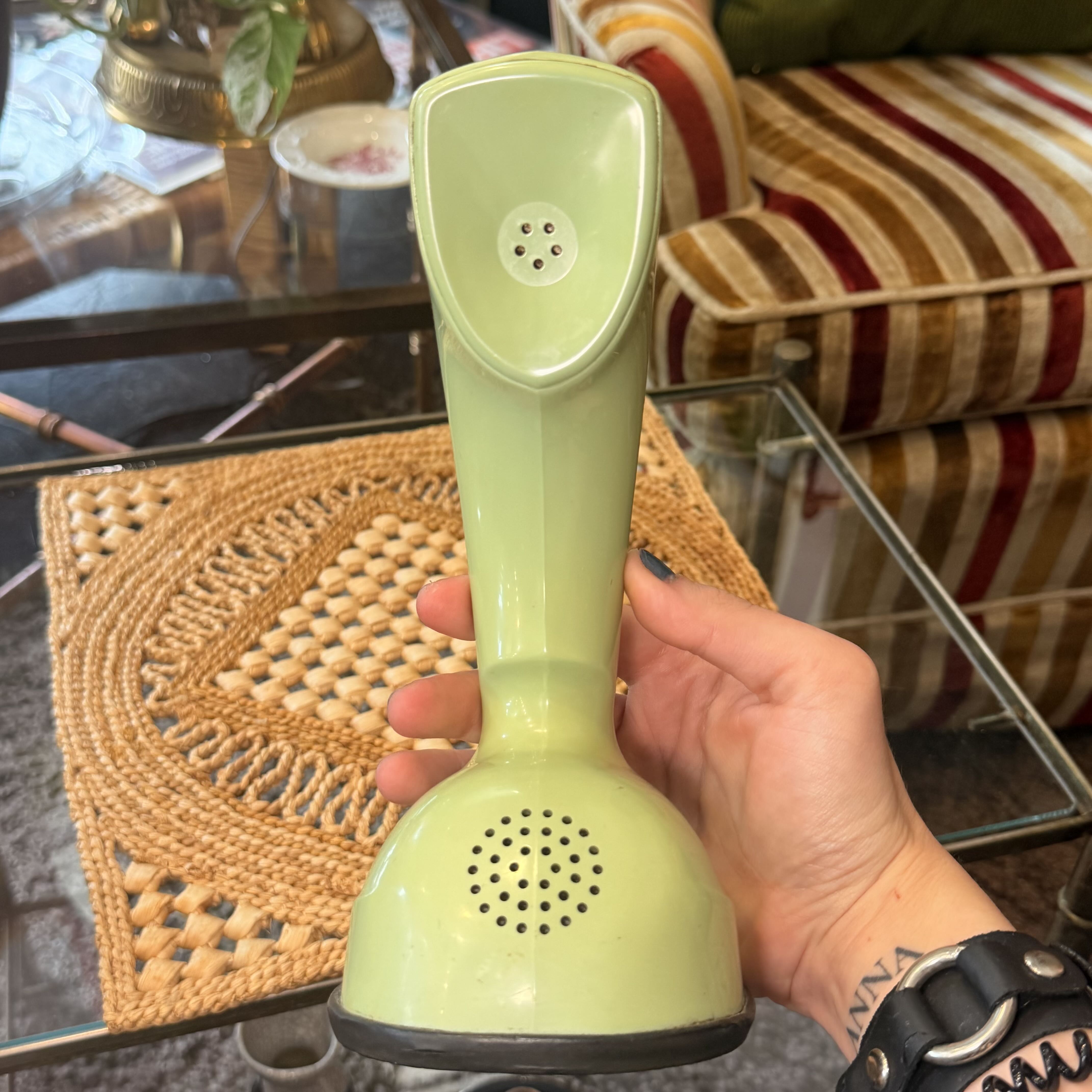 A hand holding a retro-style green cordless telephone receiver in front of a glass table and striped sofa.