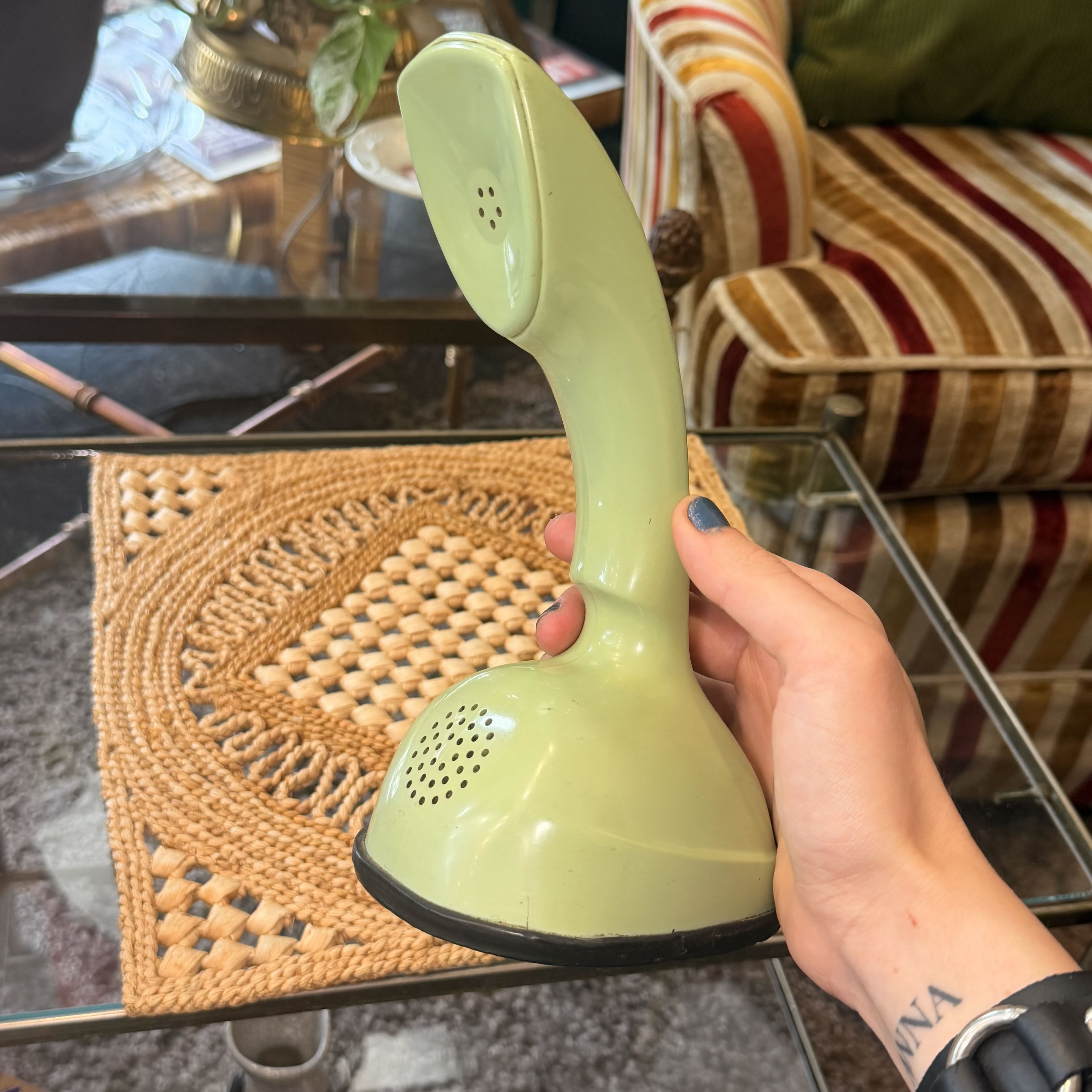 A hand holding a light green retro-style cordless telephone receiver over a glass table in a living room setting.