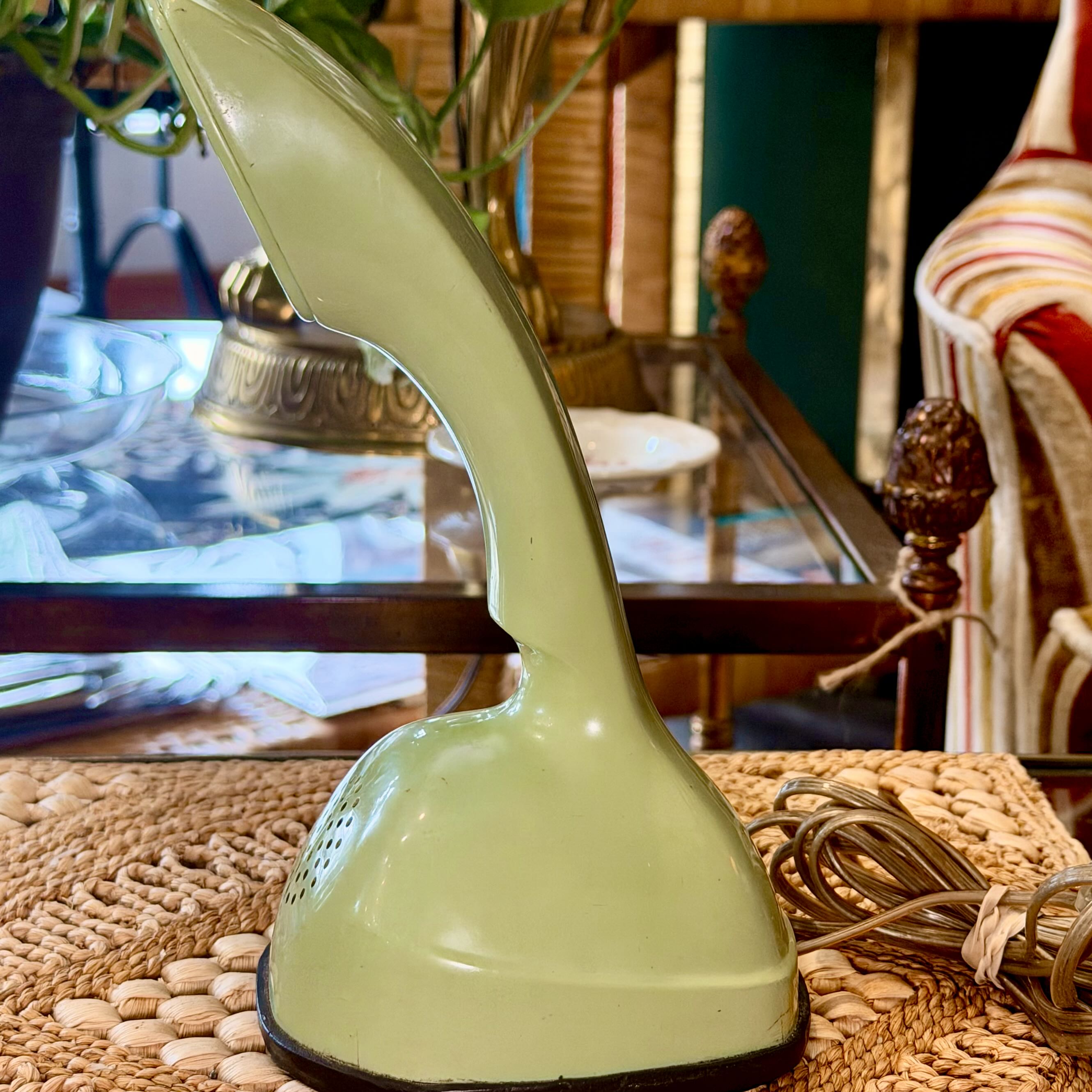 A vintage light green enamel table lamp with a curved neck and a round base, shown on a woven mat with a coiled cord in a furnished room.