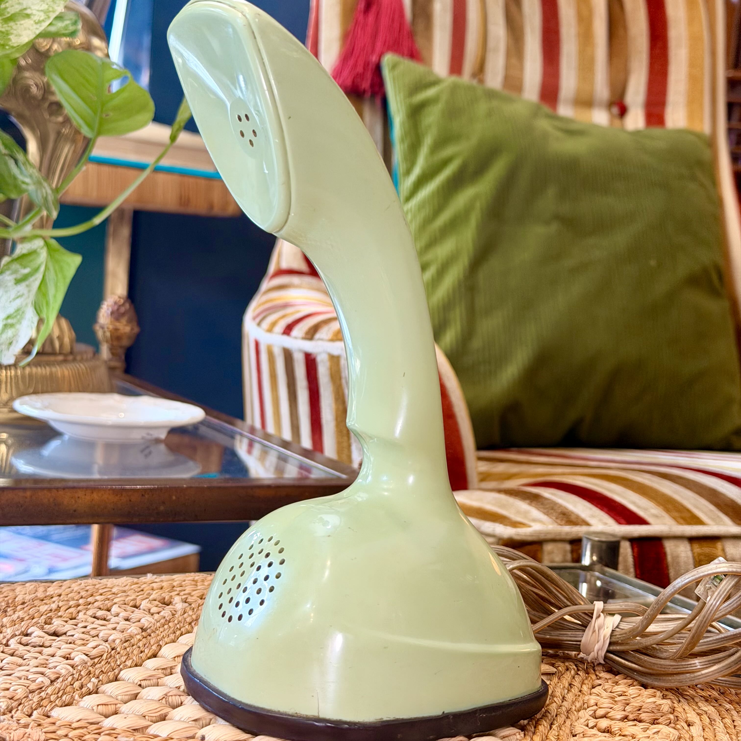 A retro-style light green rotary telephone receiver handset resting on a woven mat in a cozy indoor setting with striped upholstery, a green pillow, and a glass table in the background.