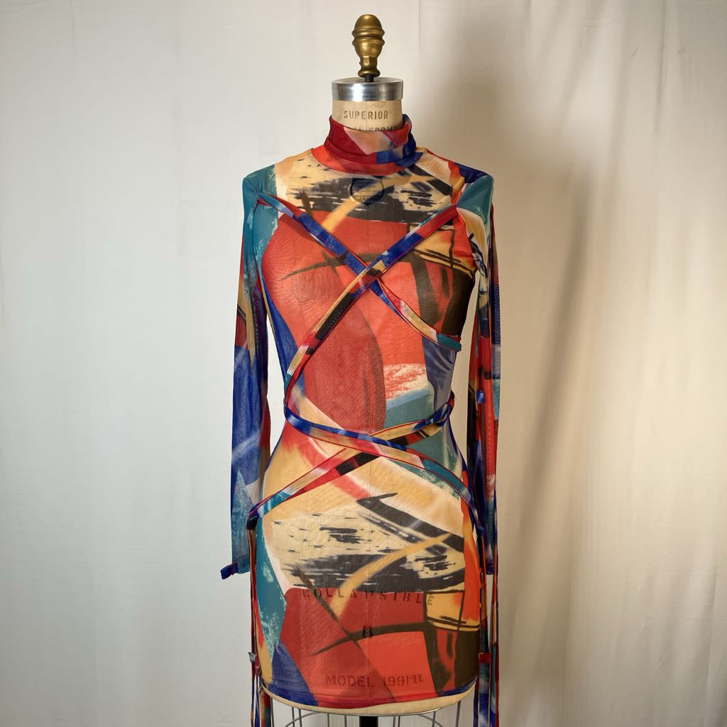 A long-sleeve mock-neck dress with an abstract geometric print, displayed on a dress form mannequin. The dress features a multicolor pattern of red, blue, beige, and black brushstrokes with criss-crossing straps across the chest and waist.