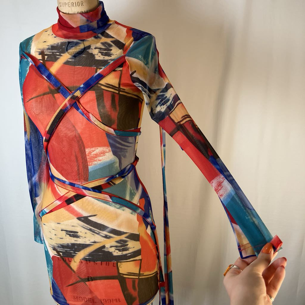 A long-sleeve sheer scarf dress with an abstract multicolor print, displayed on a dress form with a hand holding the sleeve to show its stretch, against a plain light background.