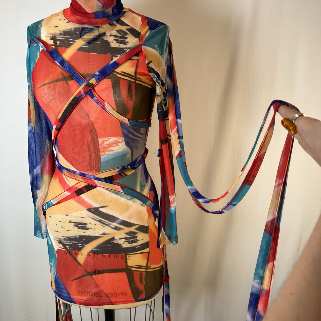 A multicolor abstract print scarf with a matching tie belt is draped over a dress form. A hand on the right holds the long, flowing tie tail.