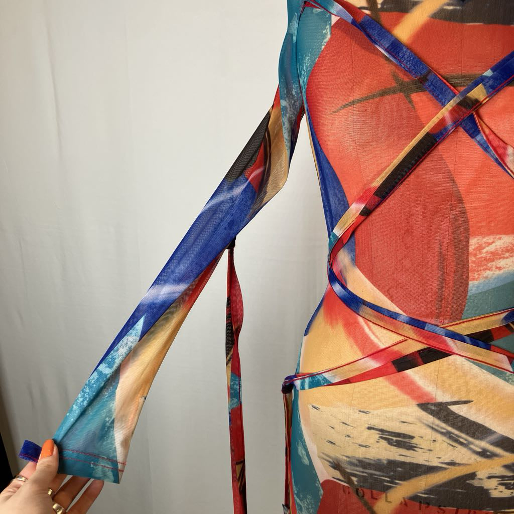 A close-up of a sheer long-sleeve wrap dress with an abstract painterly print in red, blue, beige, and black, featuring a hand holding out the sleeve to show the fabric's transparency.