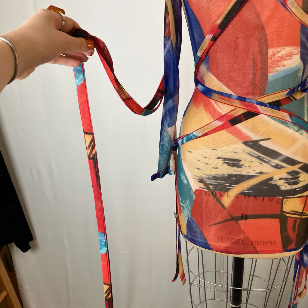 A multicolor abstract print wrap dress displayed on a wire dress form, with a hand holding the long tie straps on the left side against a white fabric backdrop.