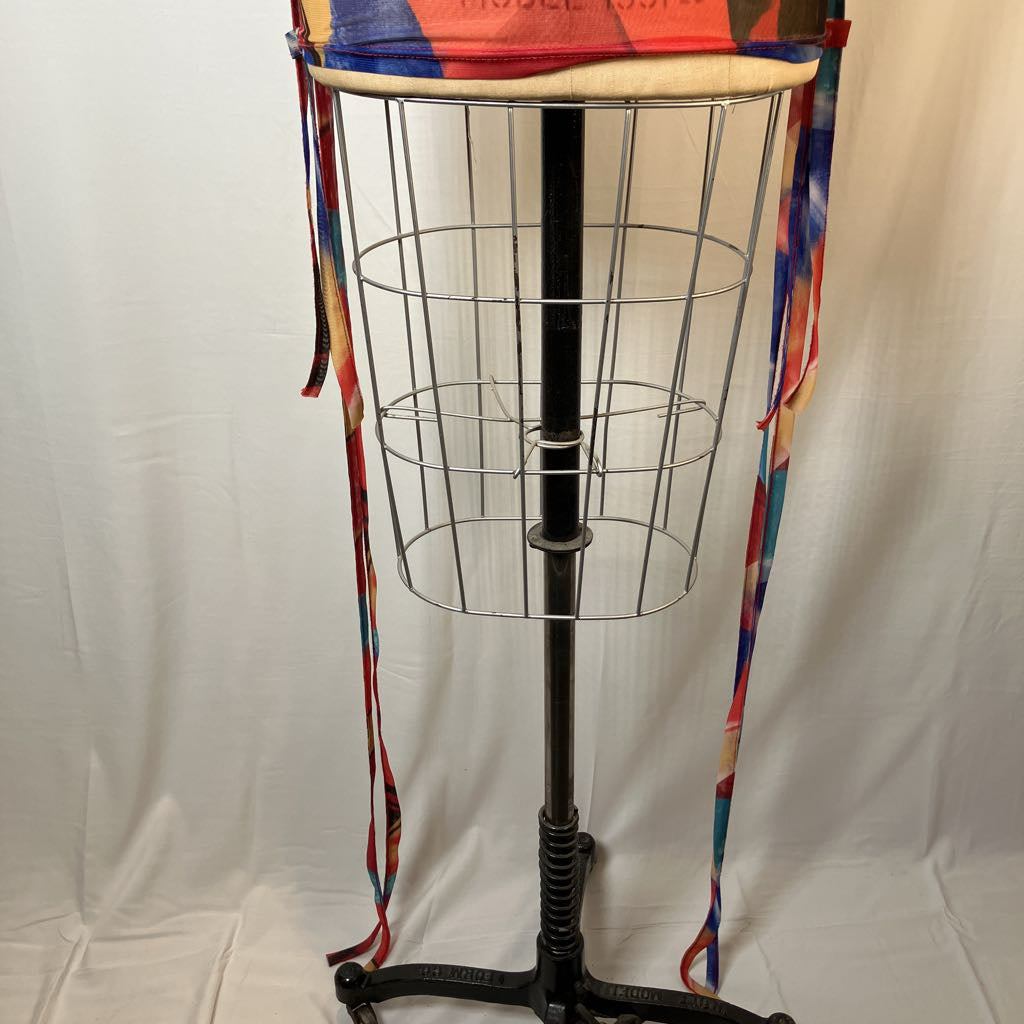 A vintage black metal garment rack stand with a wire basket cage, shown with a colorful patterned fabric hat or headband draped over the top and hanging down the sides.