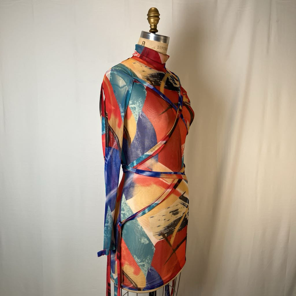 A long-sleeve abstract print top displayed on a dress form mannequin against a light fabric backdrop. The garment features a colorful brushstroke pattern in red, blue, beige, and teal with ribbon-like blue accents wrapped around the torso.
