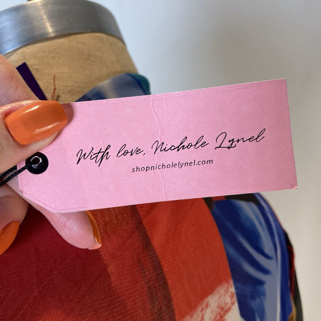 A close-up of a hand holding a pink paper brand tag that reads 'With love, Nichole Lynel' and 'shopnicholelynel.com' over a colorful garment.