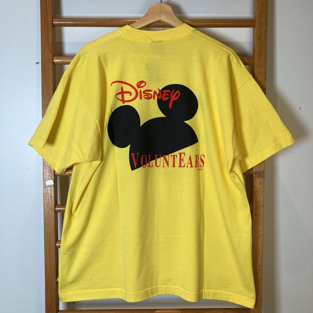 A bright yellow short-sleeve T-shirt hanging on a wooden hanger against a wooden ladder, featuring a large black Mickey Mouse silhouette on the back with the red 'Disney' logo above and 'Volunteers' text below.