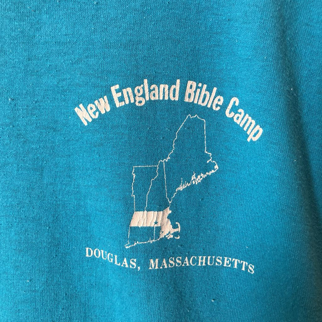 A close-up of a teal blue t-shirt featuring a vintage white graphic print with the text 'New England Bible Camp' and 'DOUGLAS, MASSASETTS' surrounding a state outline.