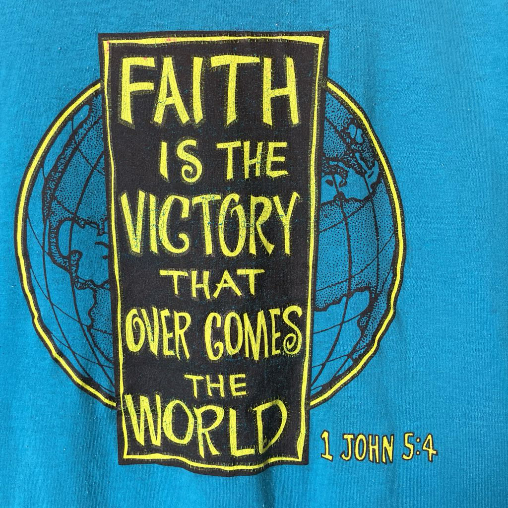 A close-up of a blue T-shirt featuring a large graphic print with a black rectangular banner outlined in yellow, reading 'FAITH IS THE VICTORY THAT OVER COMES THE WORLD,' with '1 JOHN 5:4' in yellow text at the lower right.