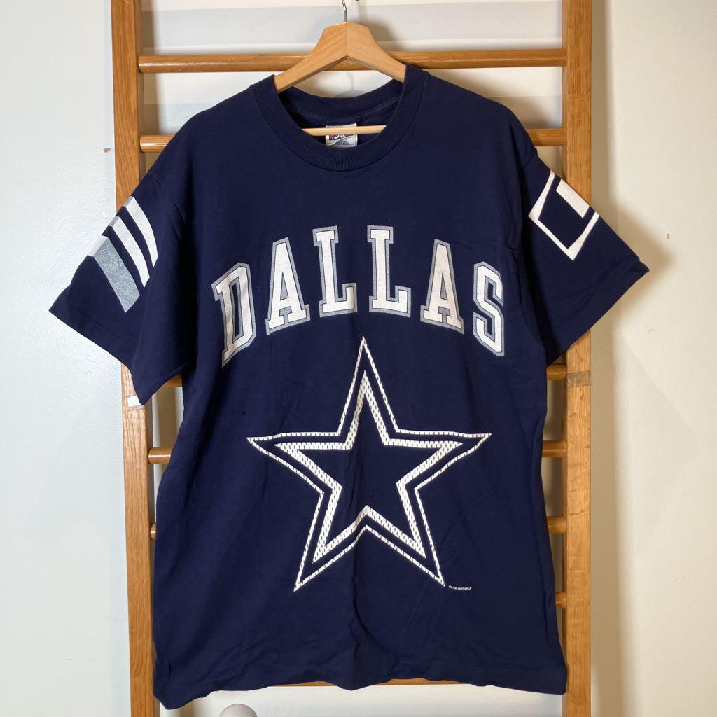 A navy blue Dallas Cowboys t-shirt hanging on a wooden hanger against a light-colored wall and wooden ladder backdrop. The shirt features 'DALLAS' in white varsity letters and the Dallas Cowboys star logo on the front, with white sleeve stripes.