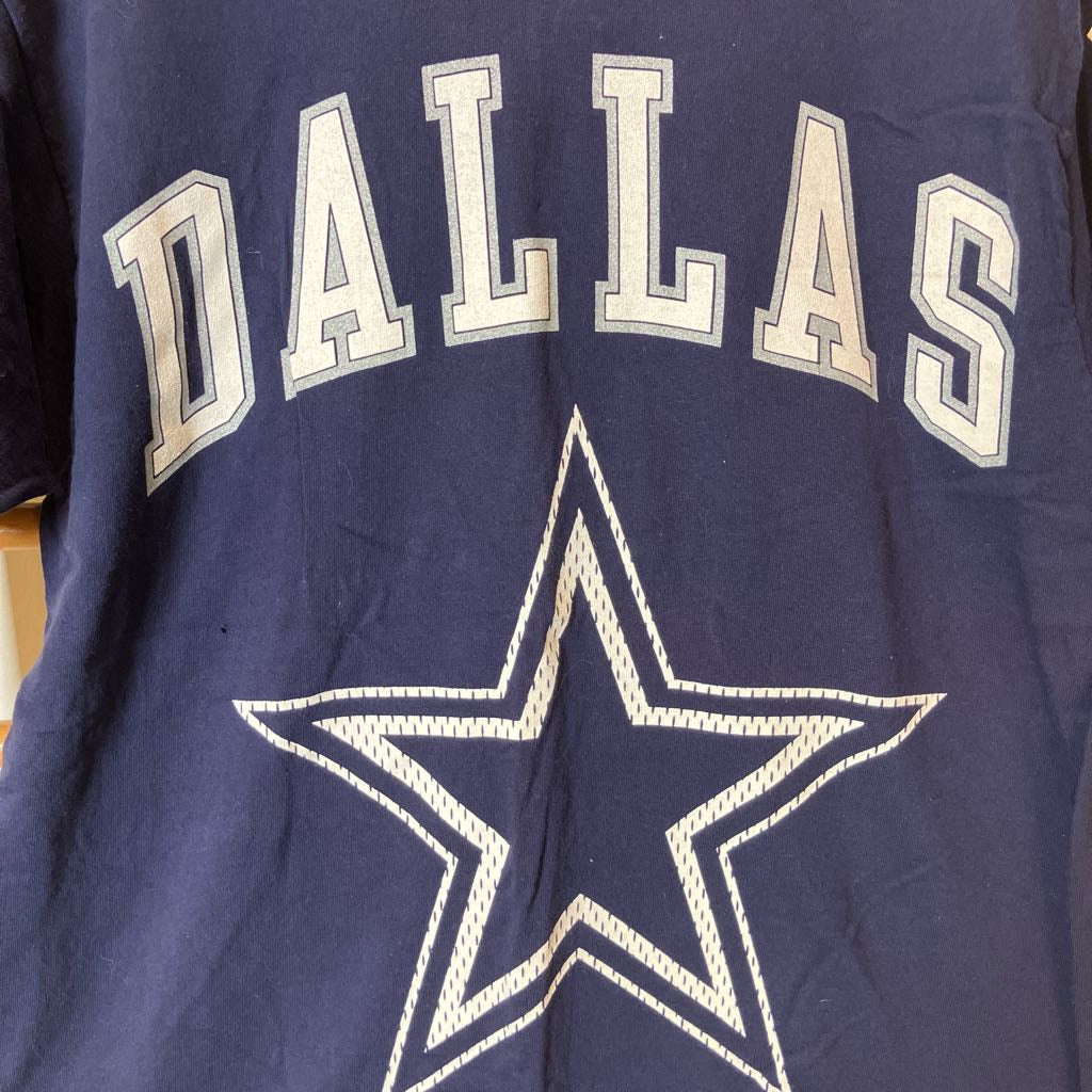 A close-up of a navy blue Dallas Cowboys t-shirt featuring the word 'DALLAS' in arched white varsity-style letters and the team's star logo below.
