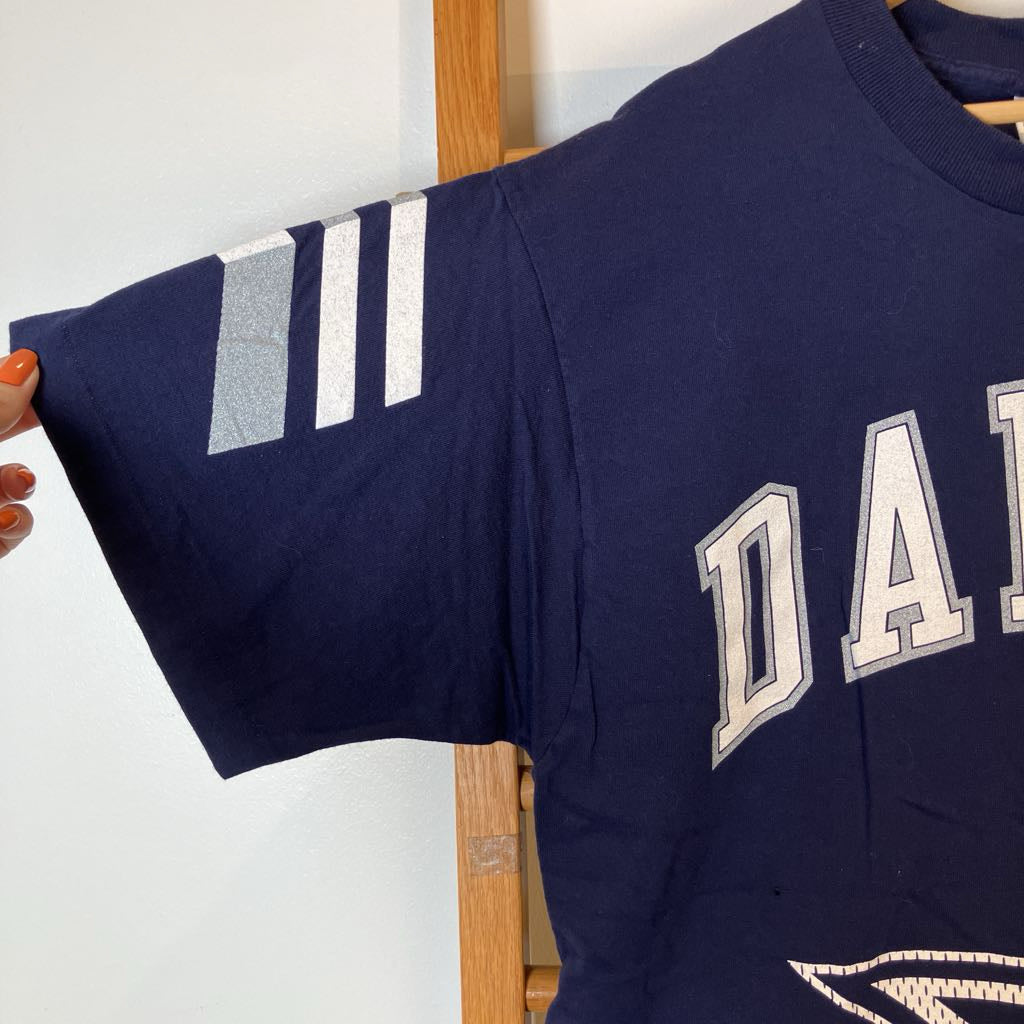 A navy blue short-sleeve t-shirt with three white stripes on the sleeve and 'DAN' printed in white varsity letters on the chest, held up by a hand.
