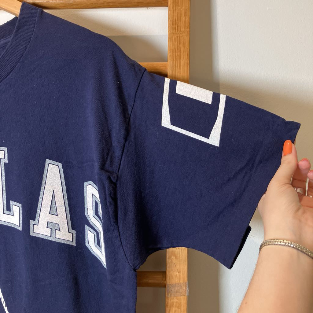 A navy blue short-sleeve t-shirt with white varsity-style lettering and a white outlined number patch on the sleeve, held up by a wooden hanger with a hand visible on the right.