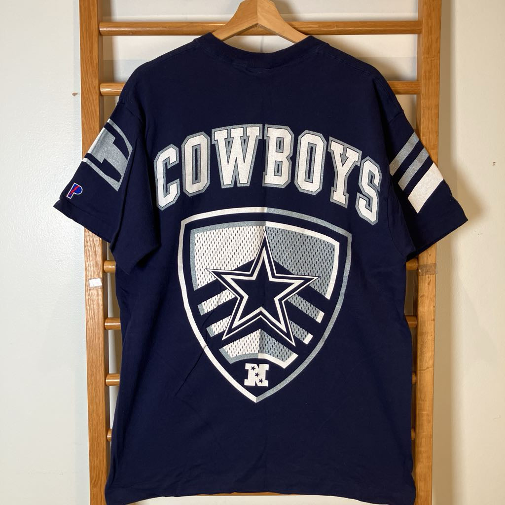 A navy blue Dallas Cowboys t-shirt hanging on a wooden hanger against a light-colored wall and wooden ladder. The back of the shirt features 'COWBOYS' in white varsity letters above a large shield logo with a silver and white star.