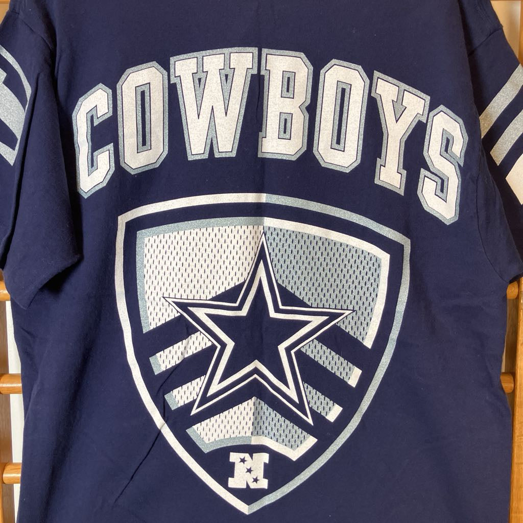 A navy blue Dallas Cowboys t-shirt featuring a large white and silver star logo and 'COWBOYS' text on the front, displayed on a wooden chair.