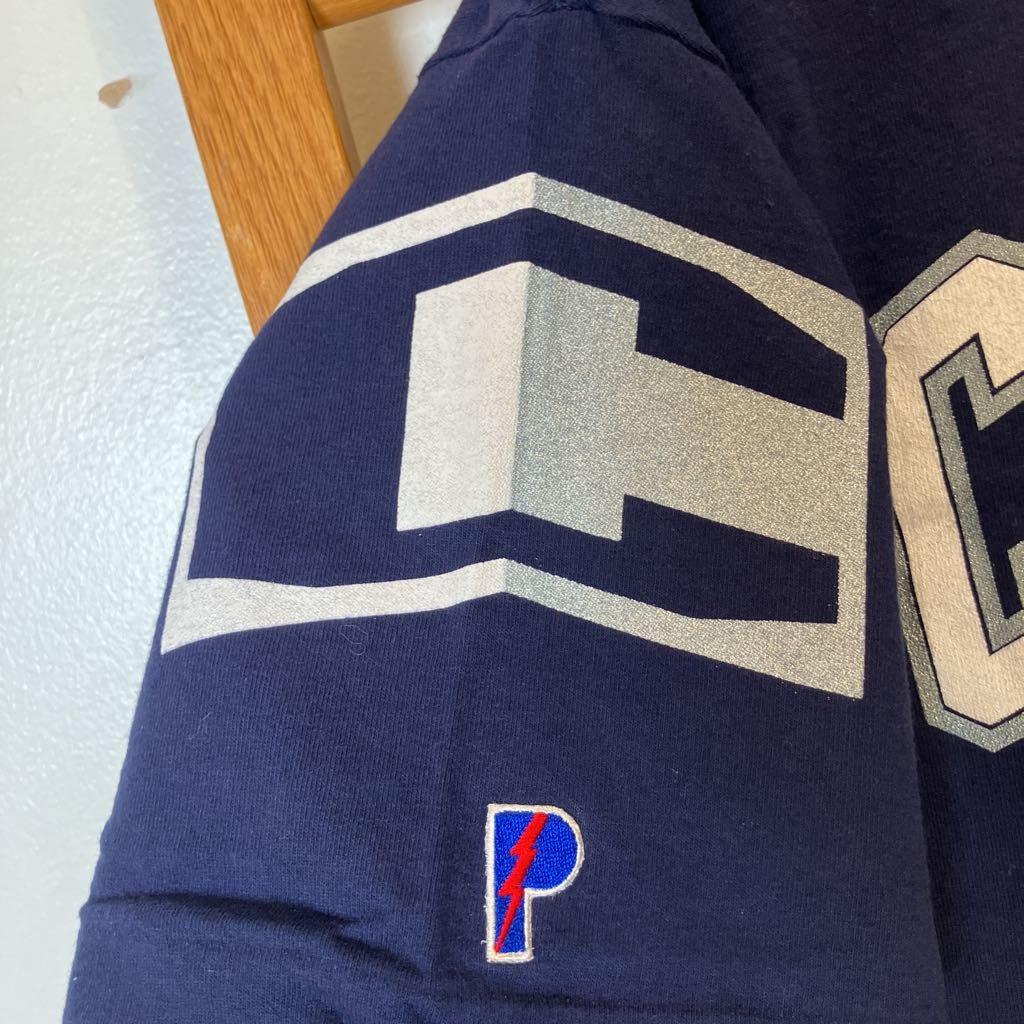 Close-up of a navy blue knit garment sleeve with a large gray and white geometric logo, a small red and blue 'P' lightning-bolt patch near the cuff, and part of a gray outlined letter visible on the adjacent sleeve.