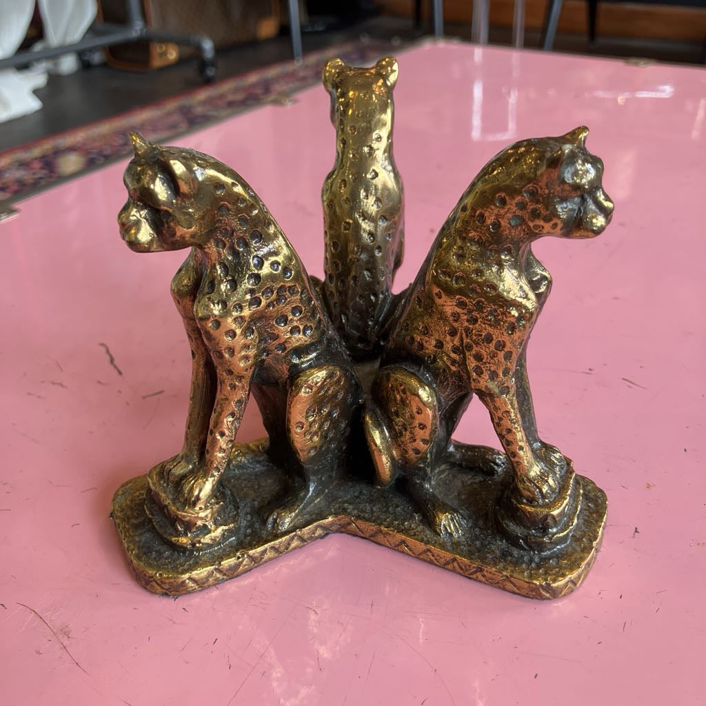 A decorative brass bookend shaped like three spotted big cats (two seated on either side, one standing in the center) on a pink tabletop.