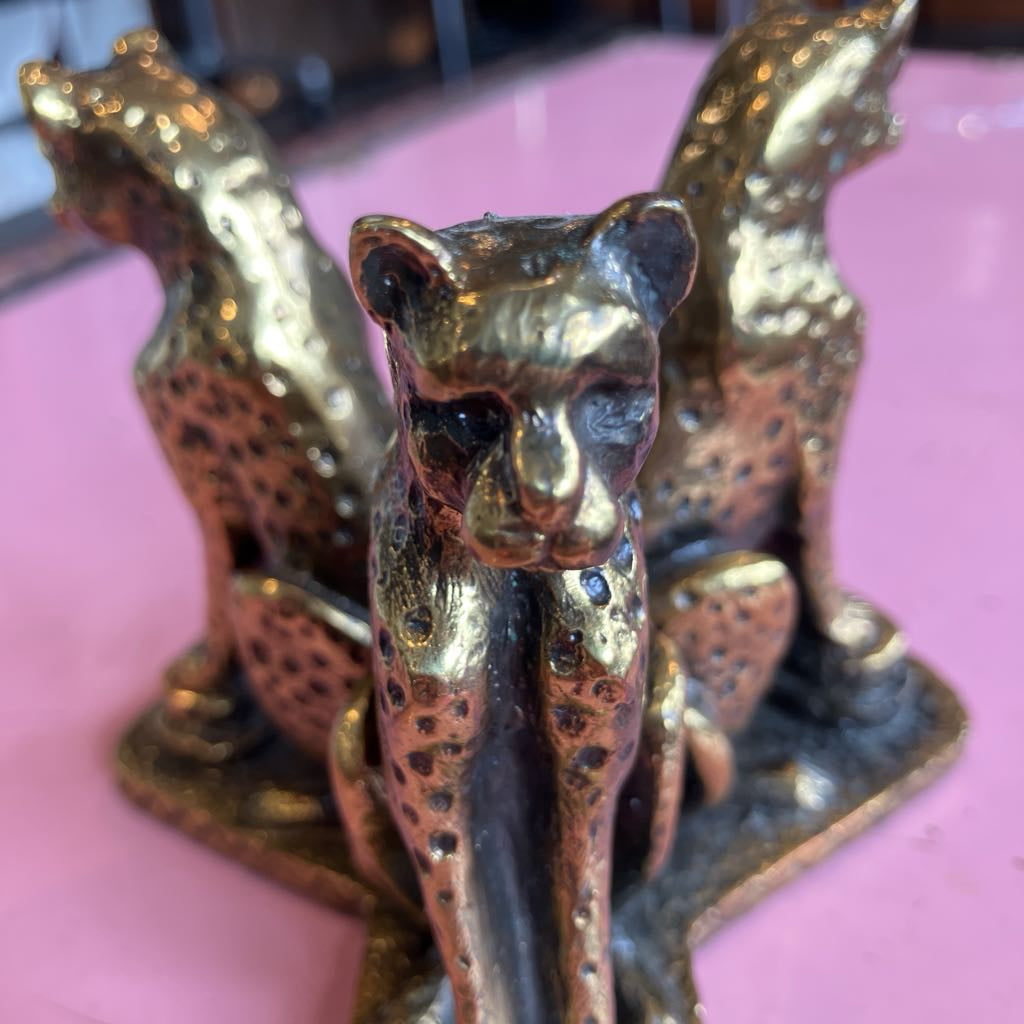 A close-up of a decorative metallic figurine featuring three spotted big cat animals, likely cheetah or leopard figures, in a bronze or gold-toned finish on a pink surface.