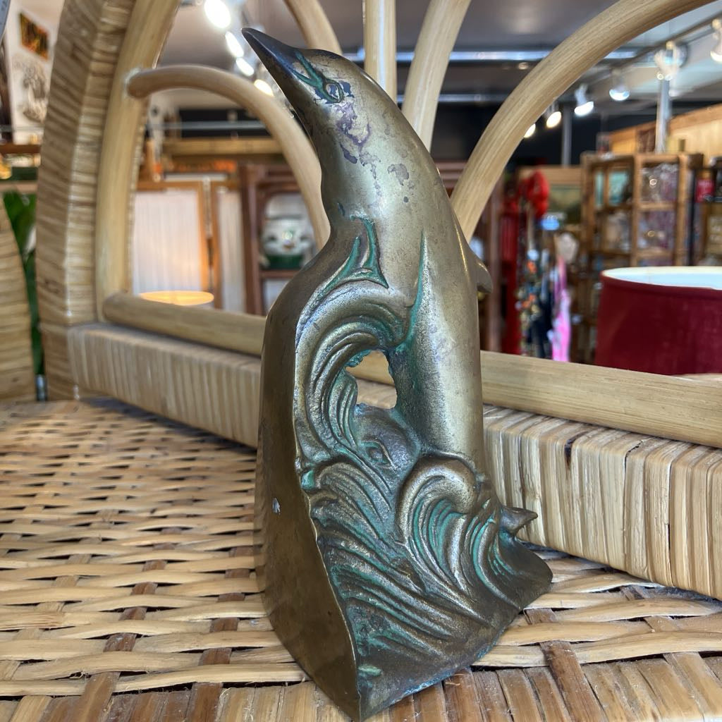 A vintage-style bronze metal dolphin figurine with wave carvings, displayed on a woven wicker surface inside a shop.