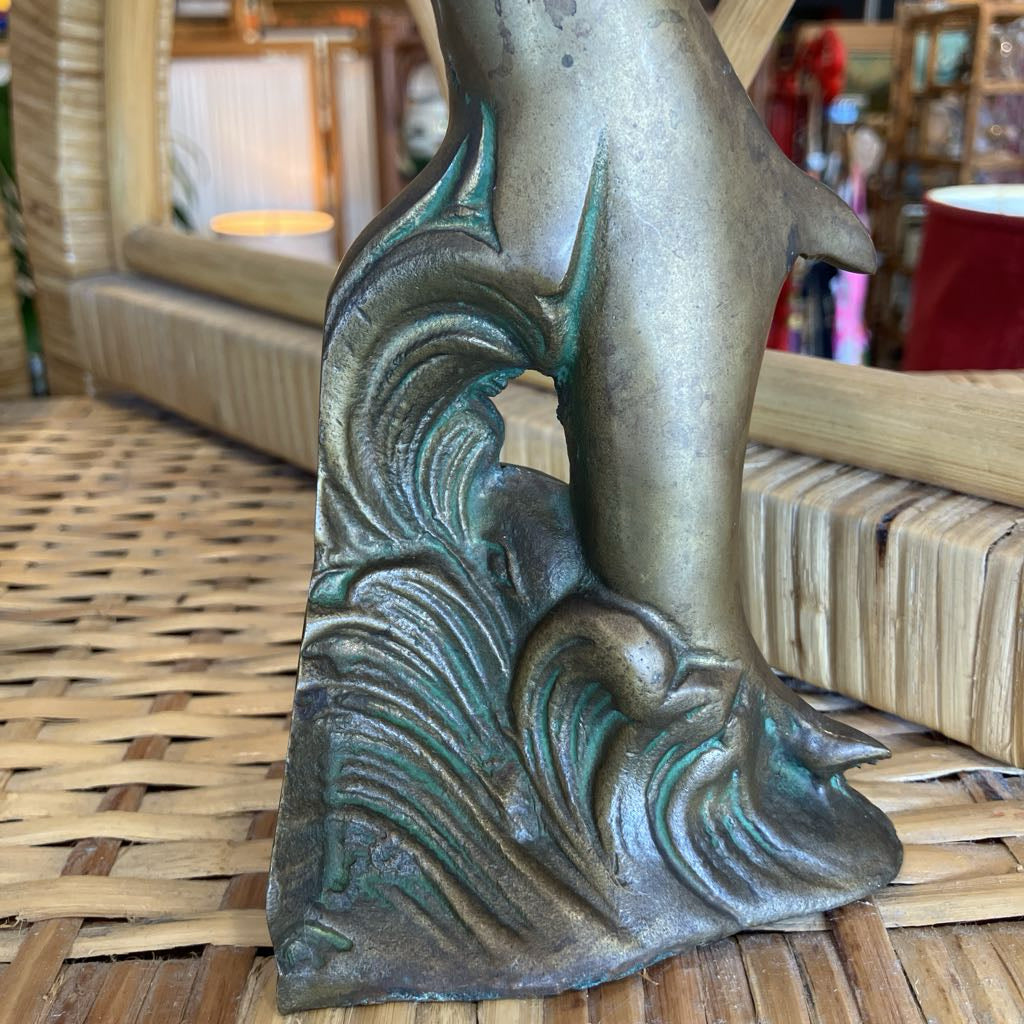 A close-up of a decorative metal sculpture featuring a stylized wave with a dolphin or shark jumping above it, displayed on a woven wicker surface in a shop setting.
