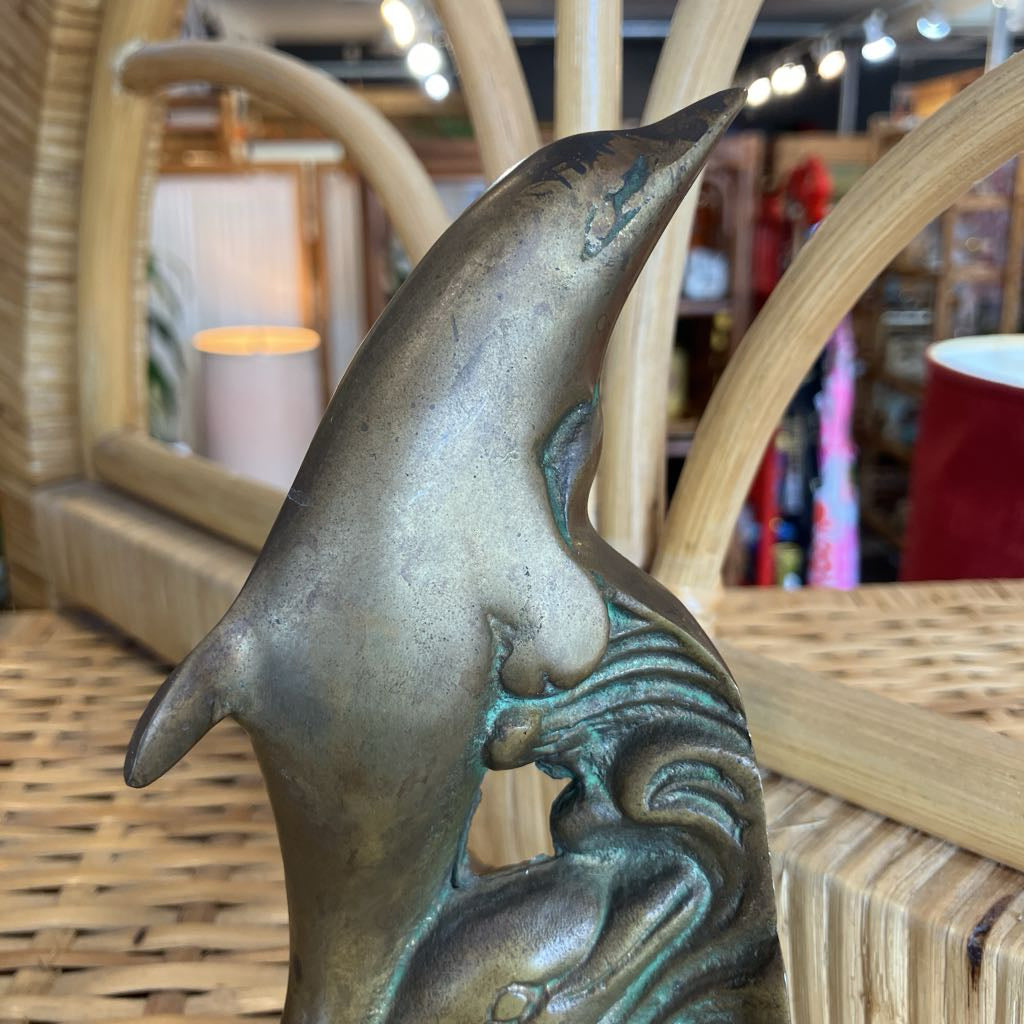 A close-up of a bronze-finished dolphin sculpture with green patina accents, resting on a woven rattan surface in a retail store setting.