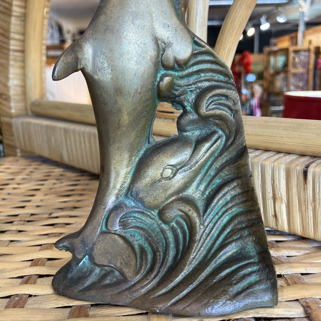A close-up of a bronze-finished decorative sculpture depicting a dolphin or whale emerging from stylized ocean waves, placed on a woven wicker surface in a shop setting.