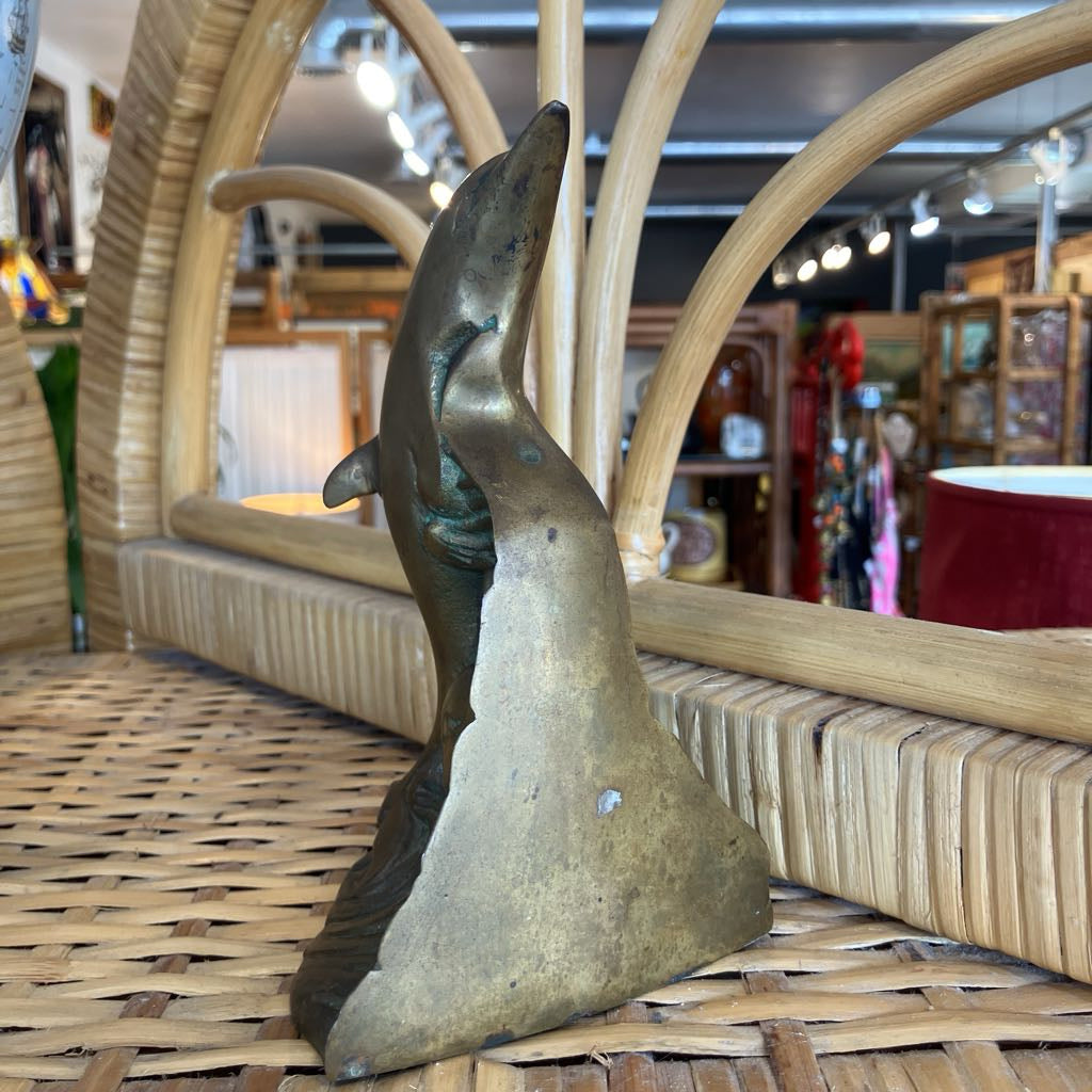 A bronze dolphin sculpture standing upright on a woven wicker surface inside a shop with wooden furniture and lighting visible in the background.