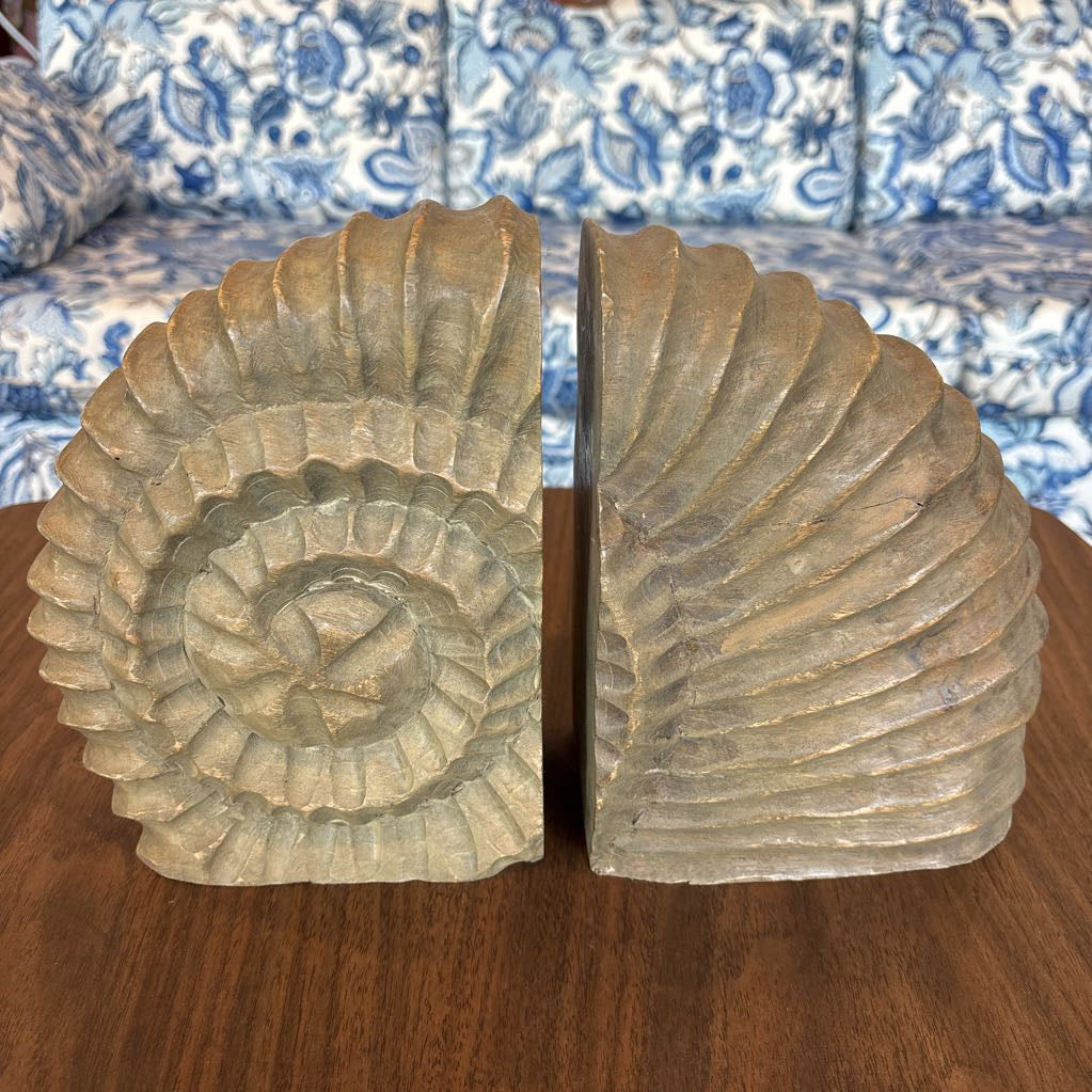 A pair of carved stone or concrete shell-shaped bookends displayed on a wooden table in front of a blue and white patterned sofa.