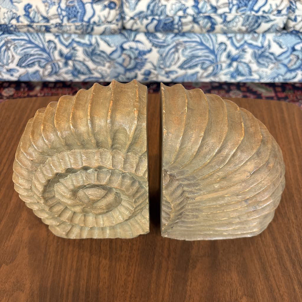 A pair of carved wooden bookends shaped like spiral ammonite shells, displayed on a wooden table with a blue and white patterned sofa in the background.
