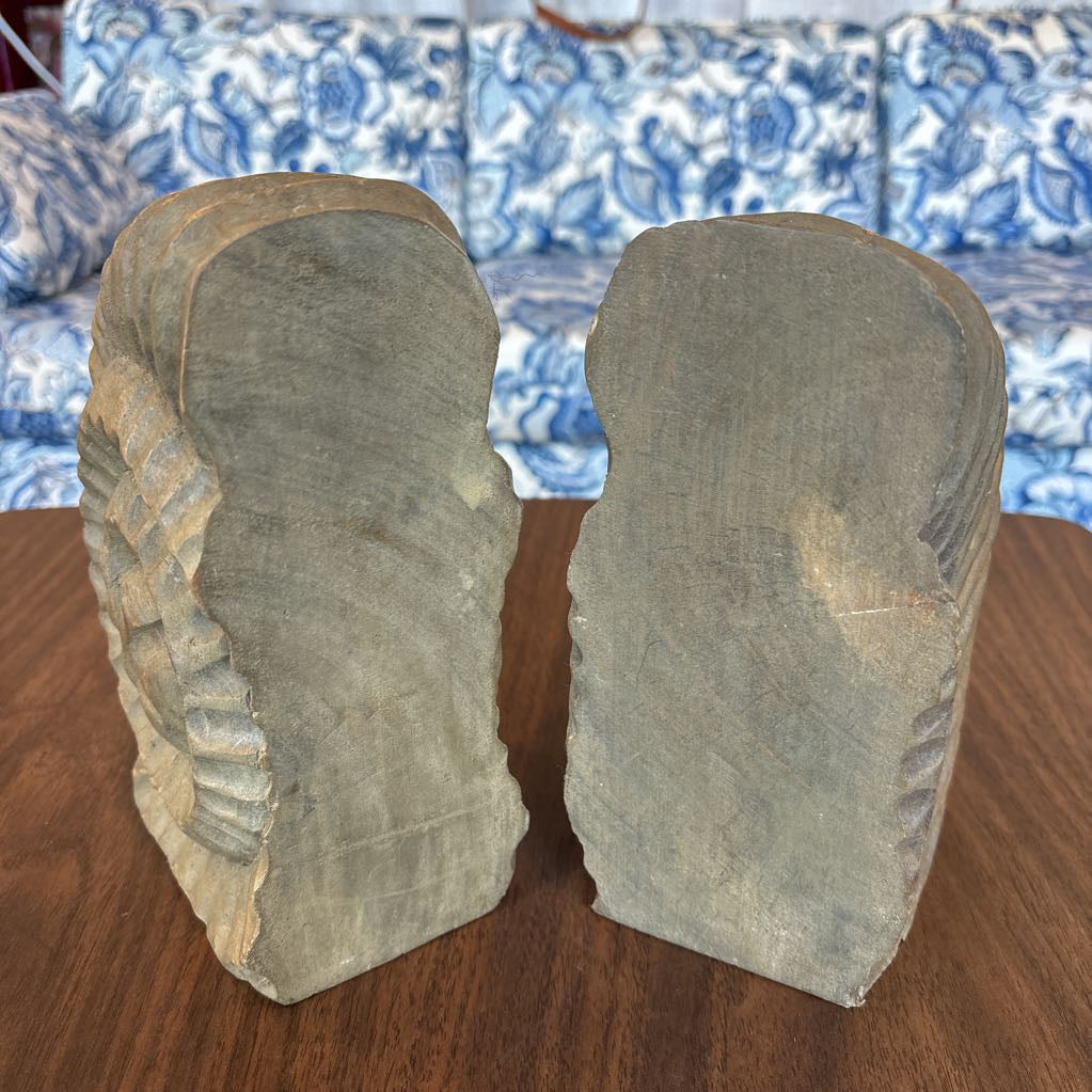 A close-up of a carved wooden bookend shaped like a seashell, showing detailed grooves and a natural wood grain finish, sitting on a wooden table with a blue and white patterned sofa in the background.
