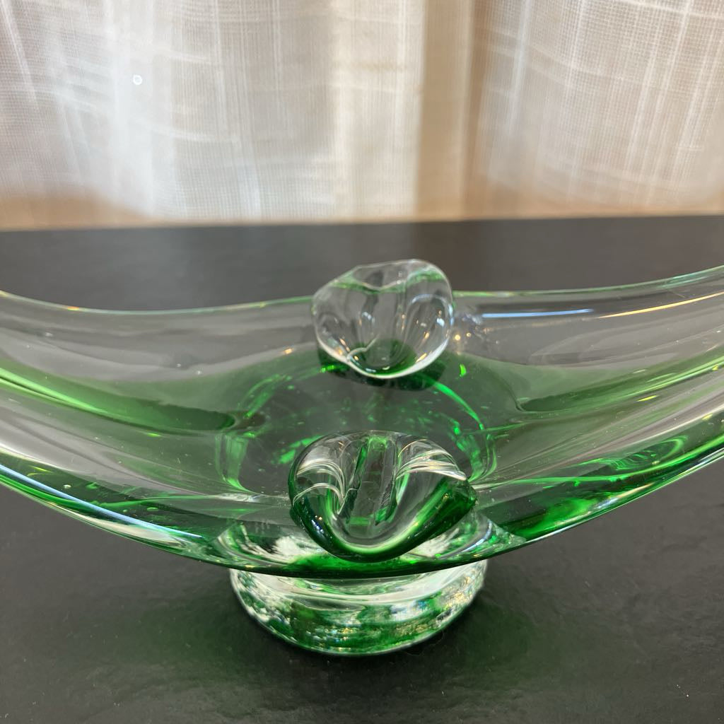A green-tinted clear glass bowl with a pedestal base and two raised decorative loops on top, photographed on a dark tabletop with a softly lit curtain in the background.