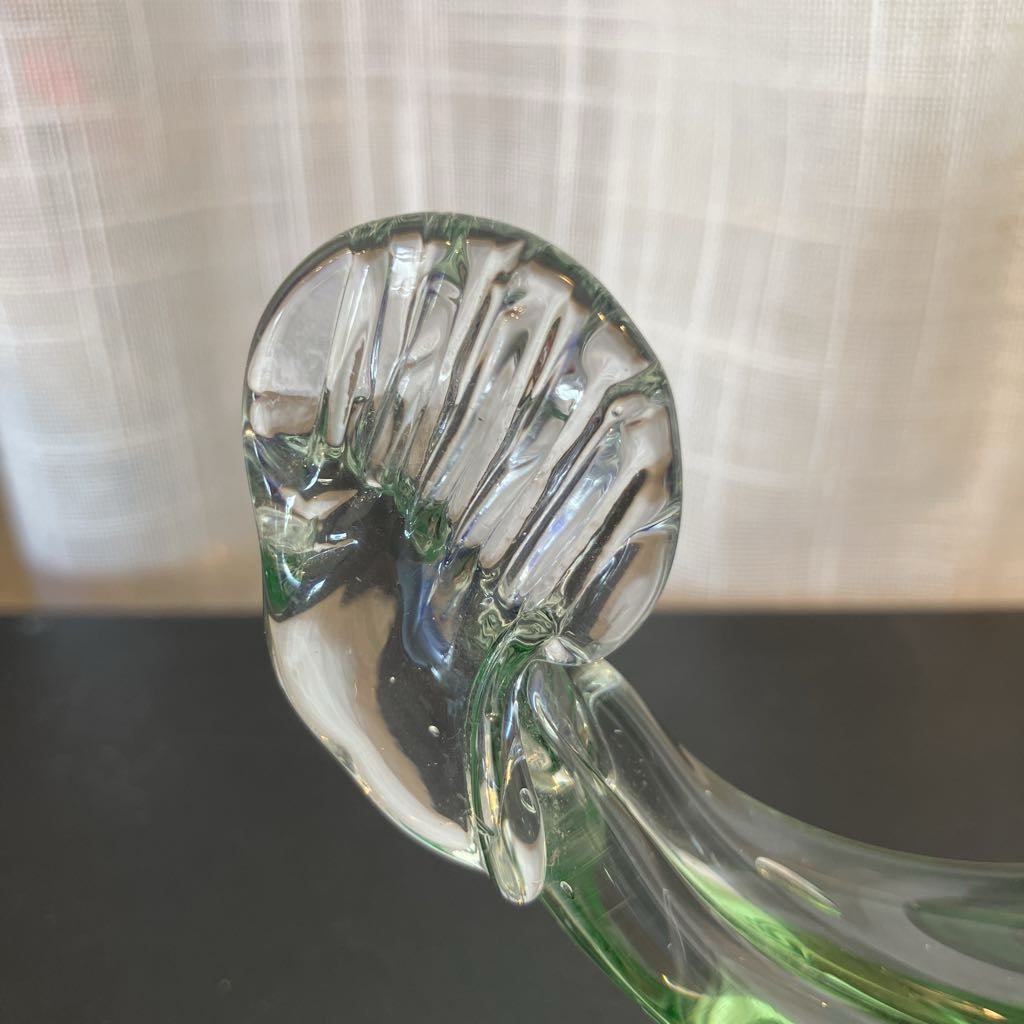 A close-up of a clear glass decorative object with a ribbed, rounded top and a curved stem, resting on a dark surface with a light curtain in the background.
