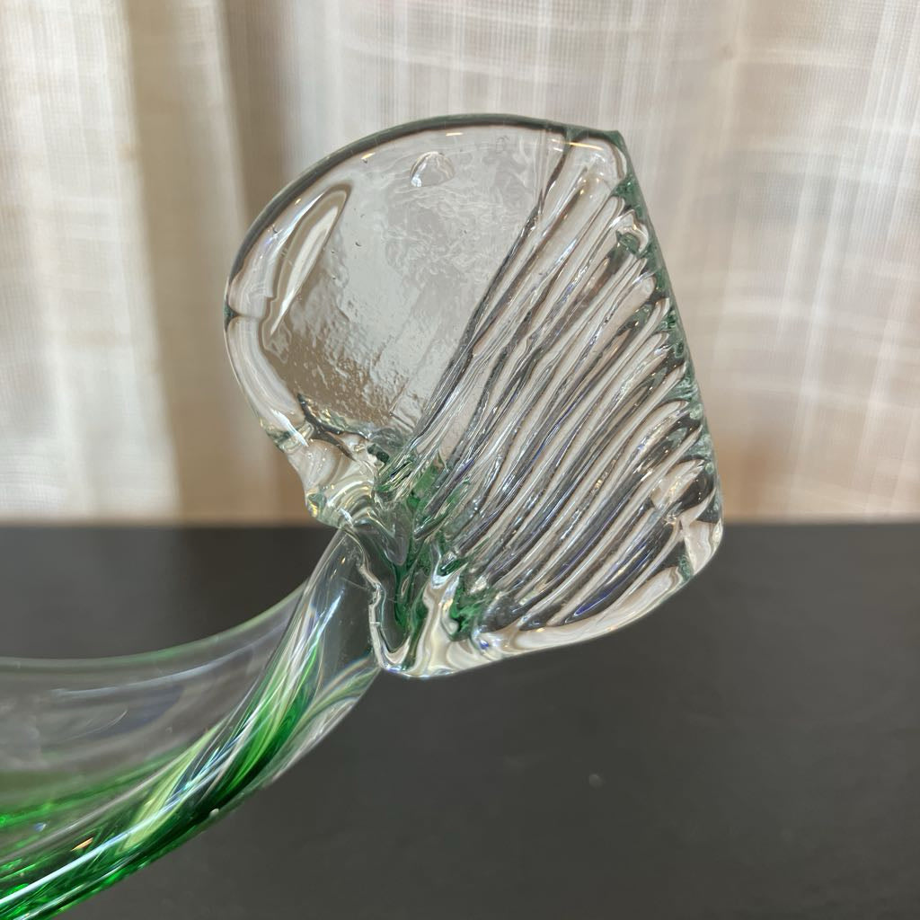 A close-up of a clear glass sculpture or decorative object with ribbed vertical texture on the upper portion and a twisted green glass stem extending into a dark surface below, set against a light curtain background.