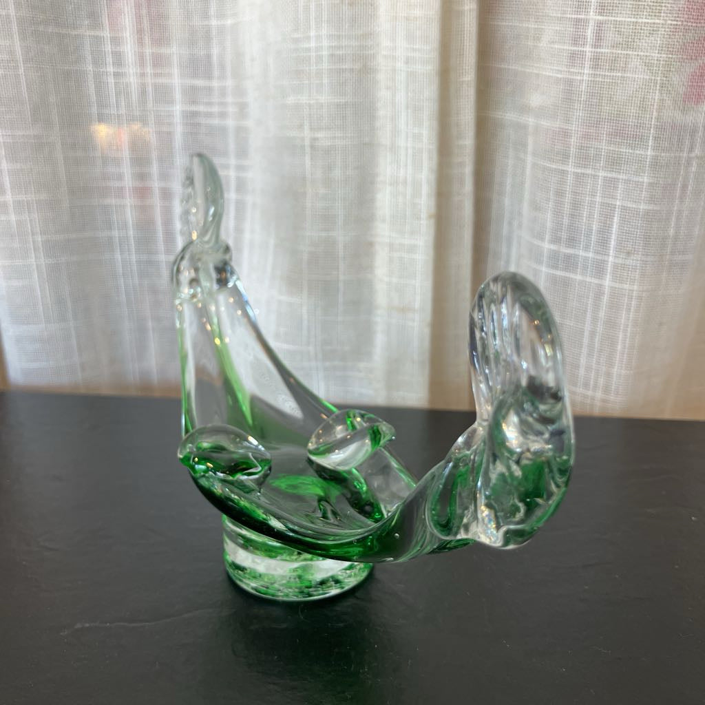 A green and clear glass decorative sculpture shaped like a stylized boat, displayed on a dark surface in front of a white curtain background.