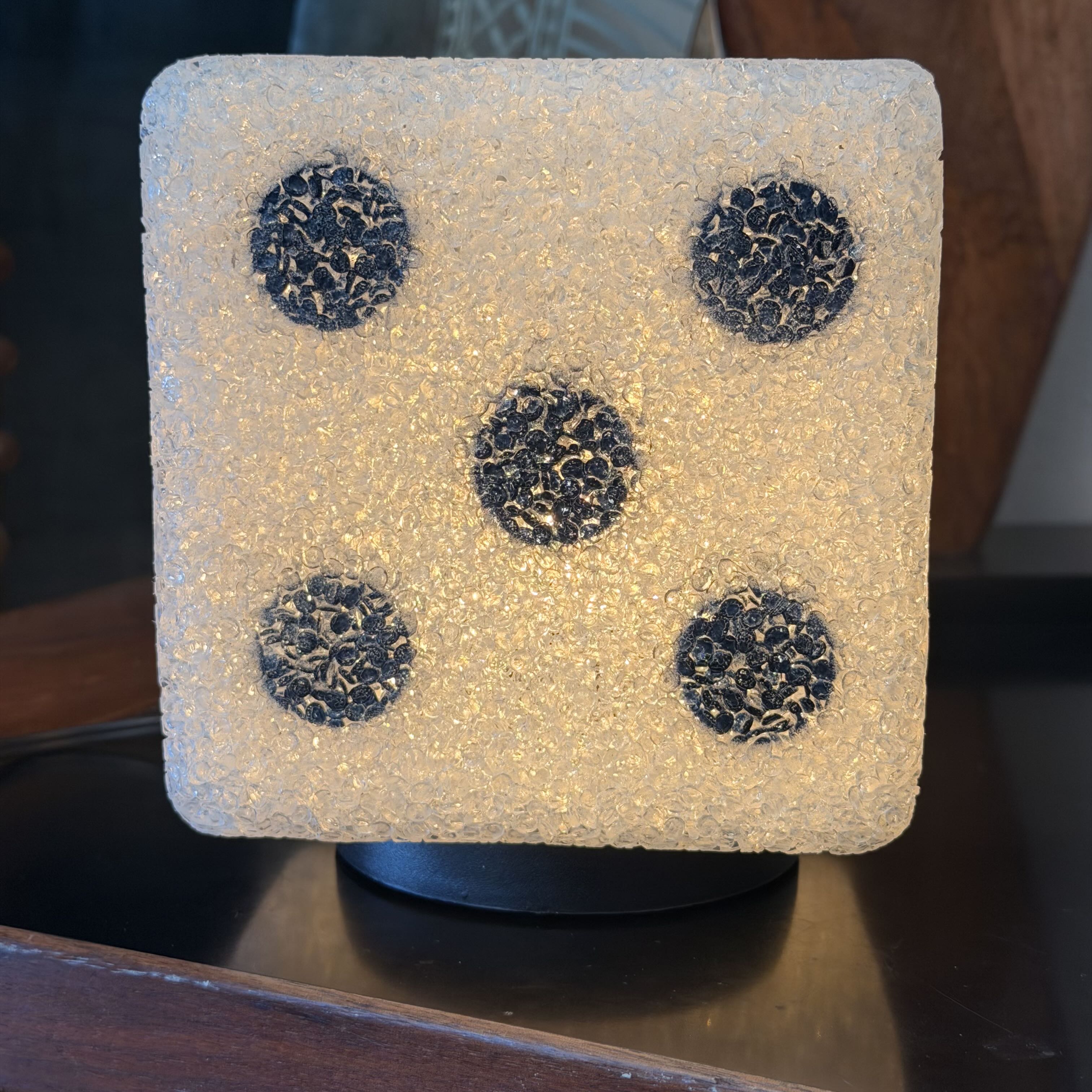 A square textured lamp or decorative light in a frosted beige finish with five black circular spots resembling a five-pawn die, shown on a dark surface.