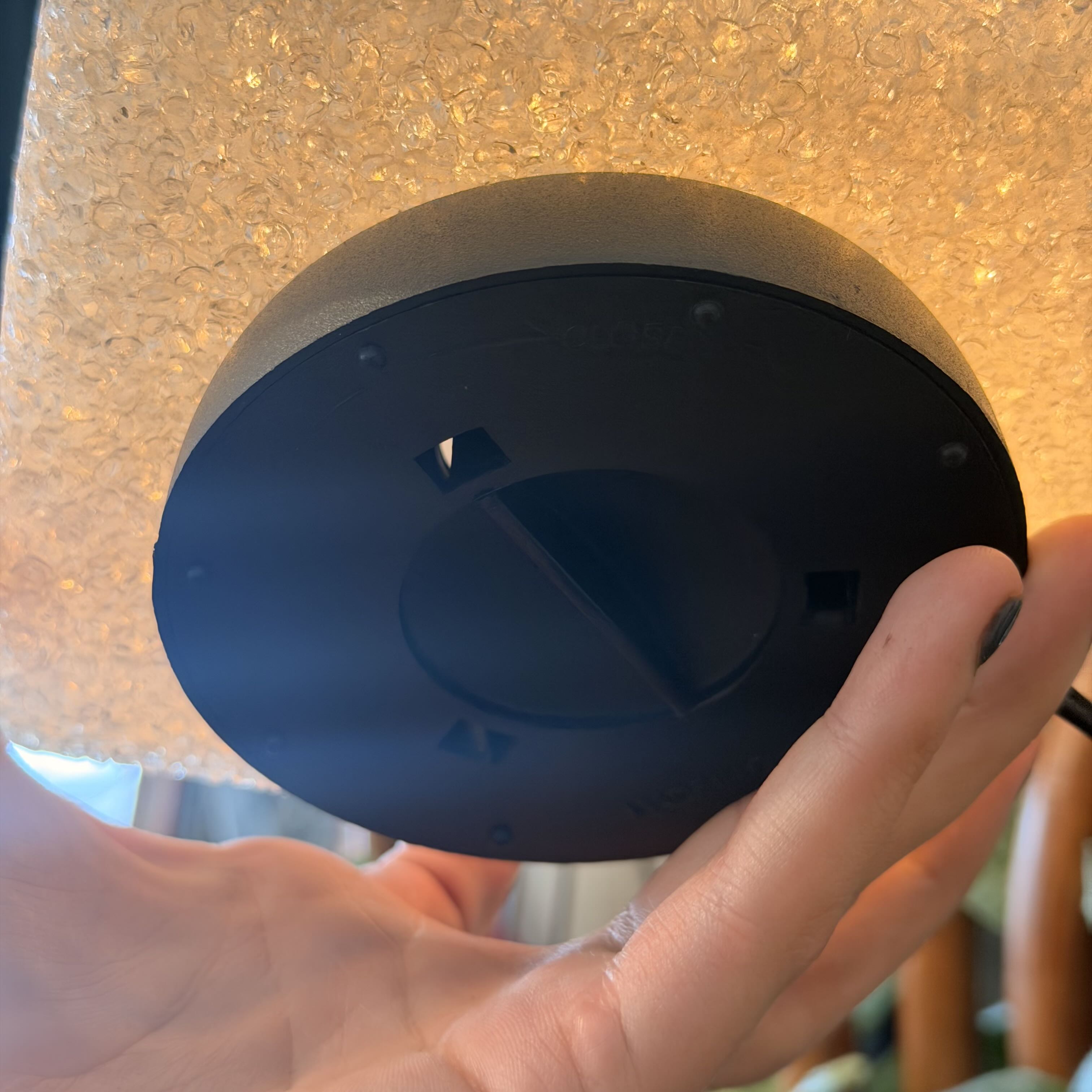 A person's hand holding a round black plastic component, likely a lamp base or socket, against a textured beige background.