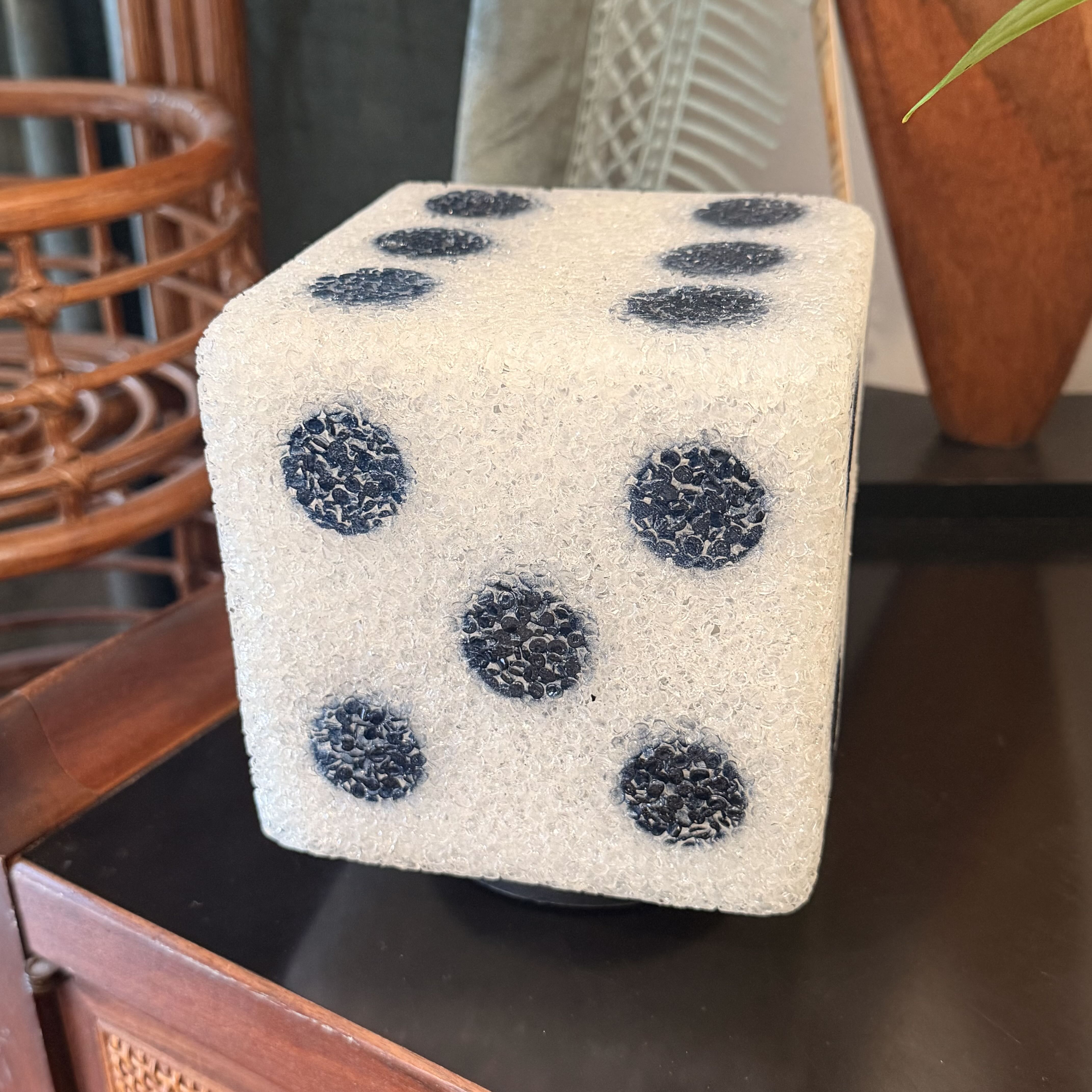 A square dice-shaped decorative object with a textured off-white surface and black dotty pattern, displayed on a dark wooden surface in an indoor setting.