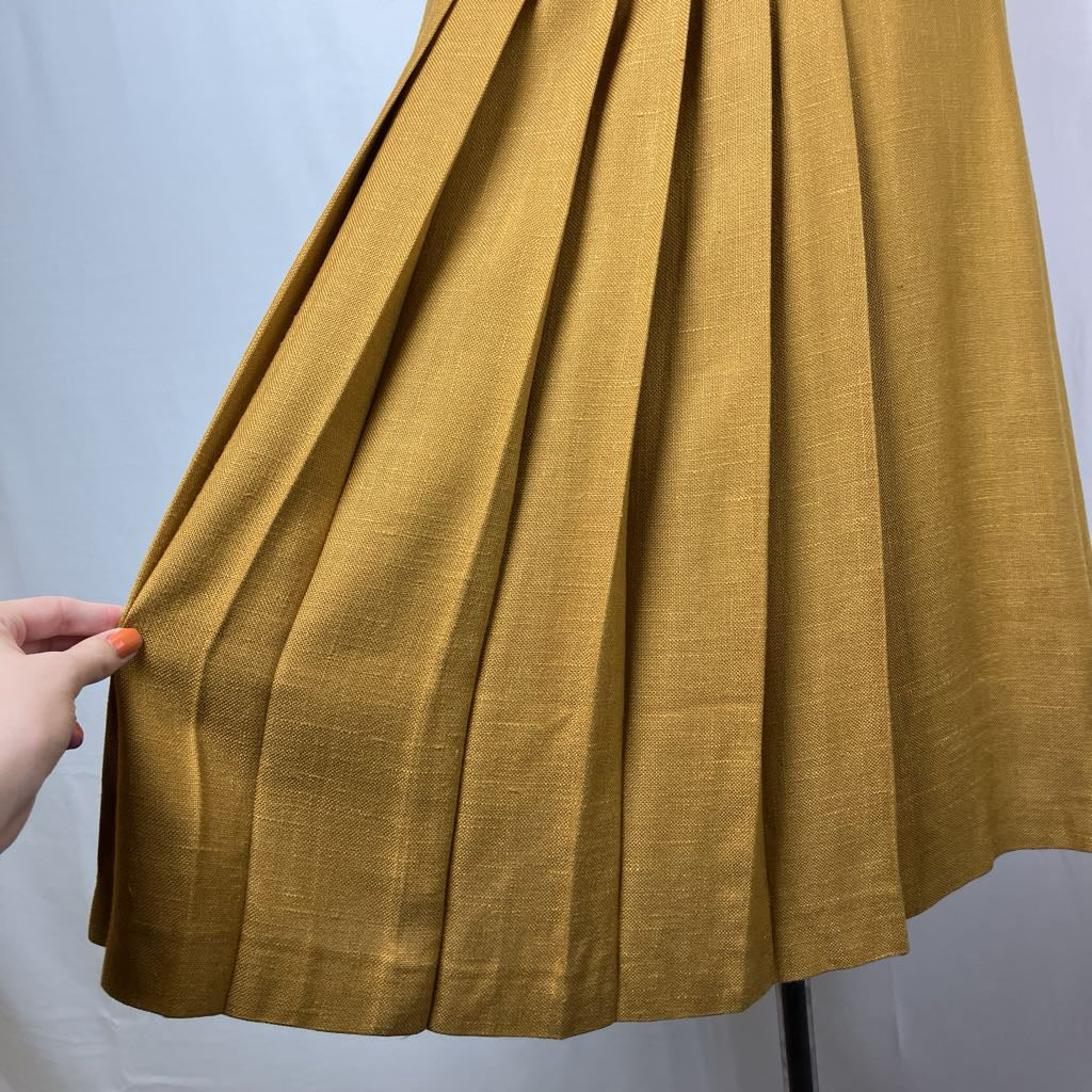 A close-up of a mustard yellow pleated skirt or drape of fabric being held by a hand, showing the textured woven material against a plain light background.