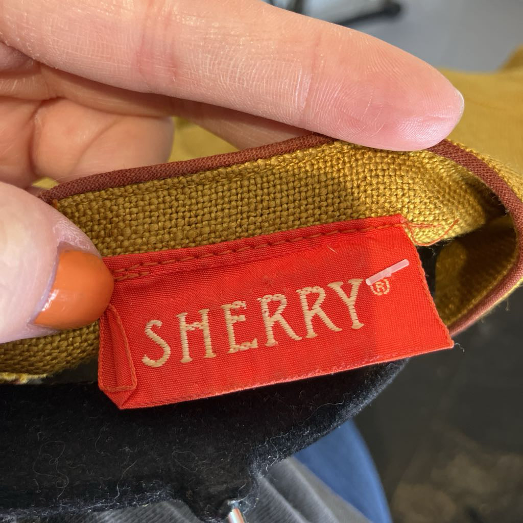 A close-up of a hand holding a mustard yellow woven fabric item, showing a red fabric brand label with 'SHERRY' embroidered in gold.