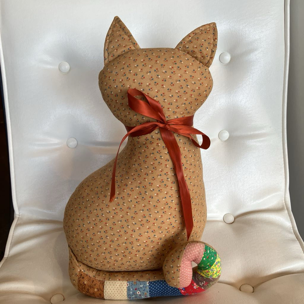 A handmade fabric cat-shaped cushion or doll shown from the back, upholstered in tan fabric with a small multicolor floral print, tied with a rust-red satin ribbon, and sitting on a white tufted chair.