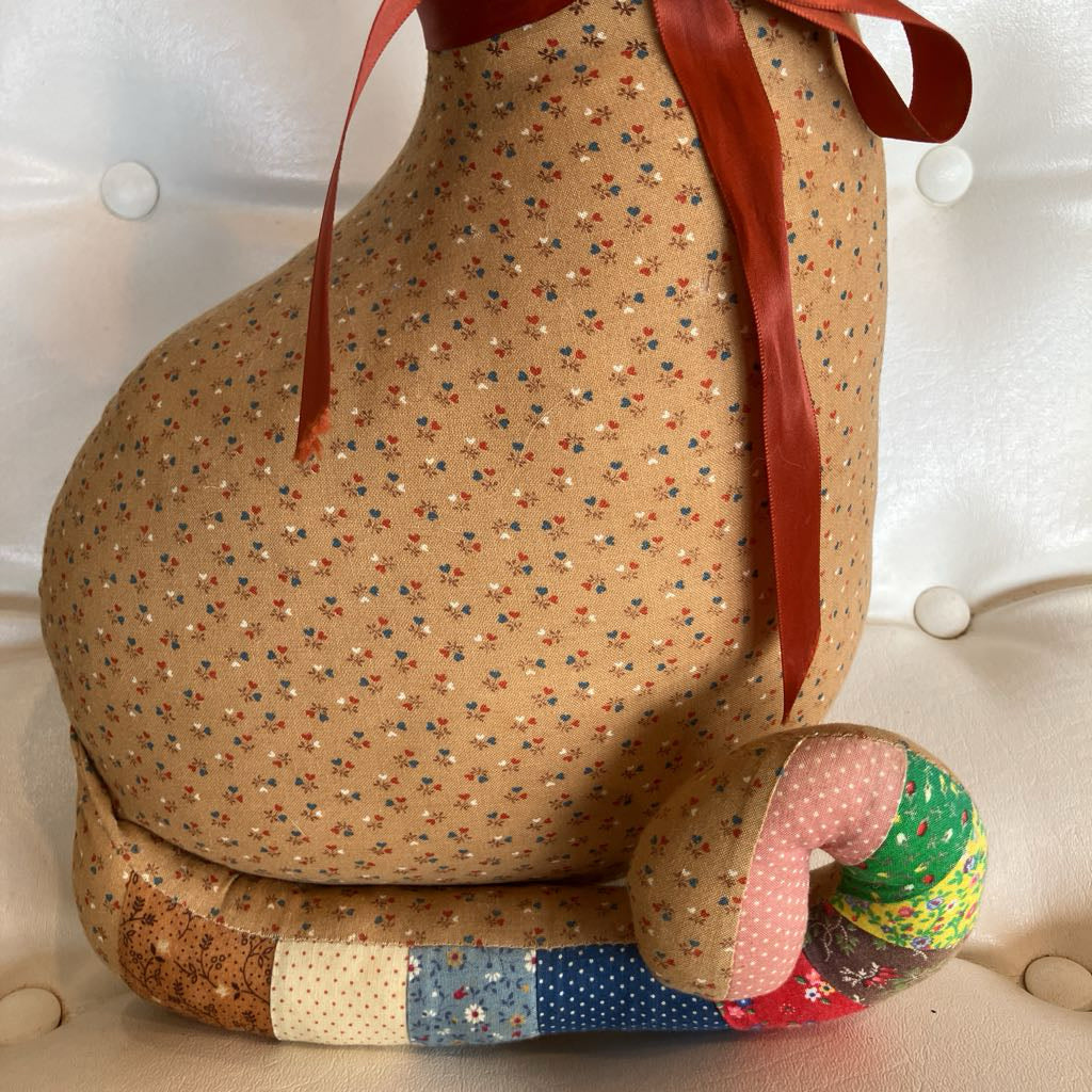 A handmade, plush-shaped cushion or pet bed in a tan fabric with a small multicolor floral print, featuring a red satin ribbon tied at the top and a patchwork-accented curved base. It is photographed on a white tufted leather chair.